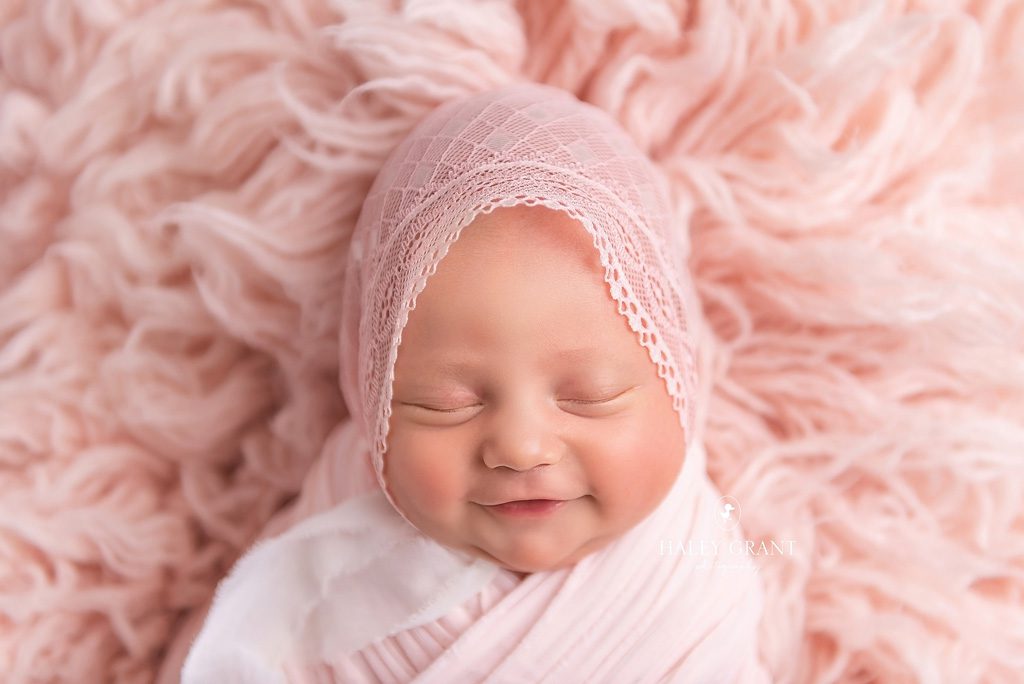 Newborn baby Isla smiling while she's swaddled in a pose at her newborn photography session. She's pink floral headband and pink wrap. She's laying on a pink backdrop. All props are provided by Haley. Photo taken at Haley Grant Photography, Cedar Park Texas Newborn Portrait studio.