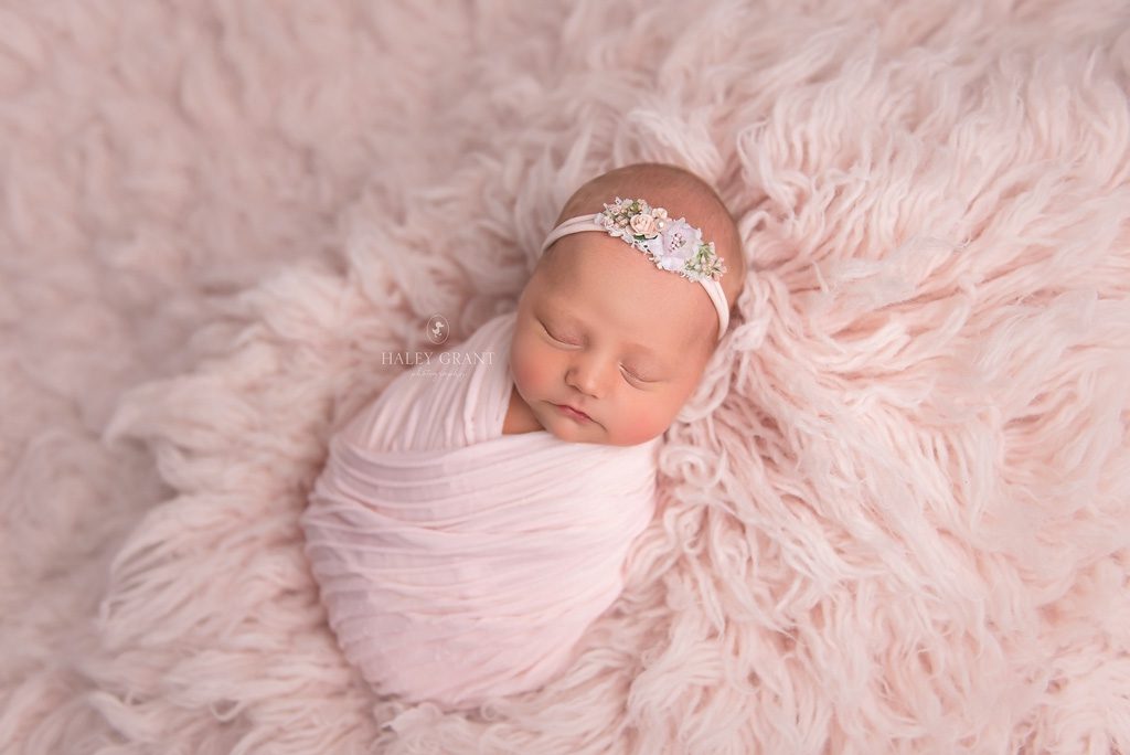 Newborn baby Isla swaddled in a pose at her newborn photography session. She's pink floral headband and pink wrap. She's laying on a pink backdrop. All props are provided by Haley. Photo taken at Haley Grant Photography, Cedar Park Texas Newborn Portrait studio.