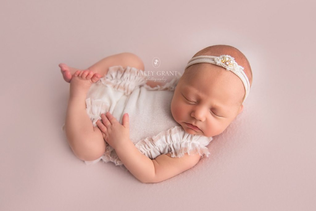 Newborn baby Isla laying in a pose at her newborn photography session. She's wearing a white pearl headband and white outfit. She's laying on a pink backdrop. All props are provided by Haley. Photo taken at Haley Grant Photography, Cedar Park Texas Newborn Portrait studio.