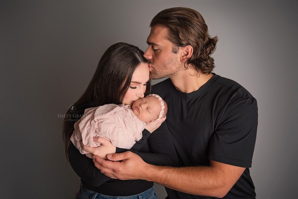New parents holding their newborn baby girl Isla and kissing each other sweetly. Photo taken at Haley Grant Photography, Cedar Park Texas Newborn Portrait studio.