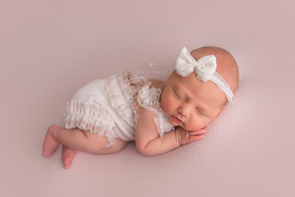 Newborn baby Isla laying in a pose at her newborn photography session. She's wearing a white bow and white outfit. She's laying on a pink backdrop. All props are provided by Haley. Photo taken at Haley Grant Photography, Cedar Park Texas Newborn Portrait studio.